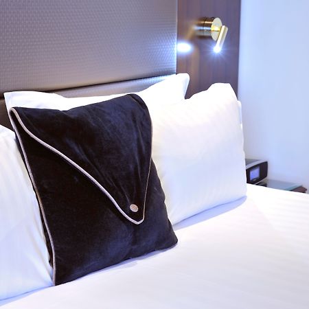 墨尔本斯坦福广场酒店 外观 照片 The photo shows a neatly arranged bed with white linens. There are several pillows, with one black pillow featuring a distinctive design on top of the white pillows. The headboard of the bed is visible, along with a stylish wall-mounted light fixture