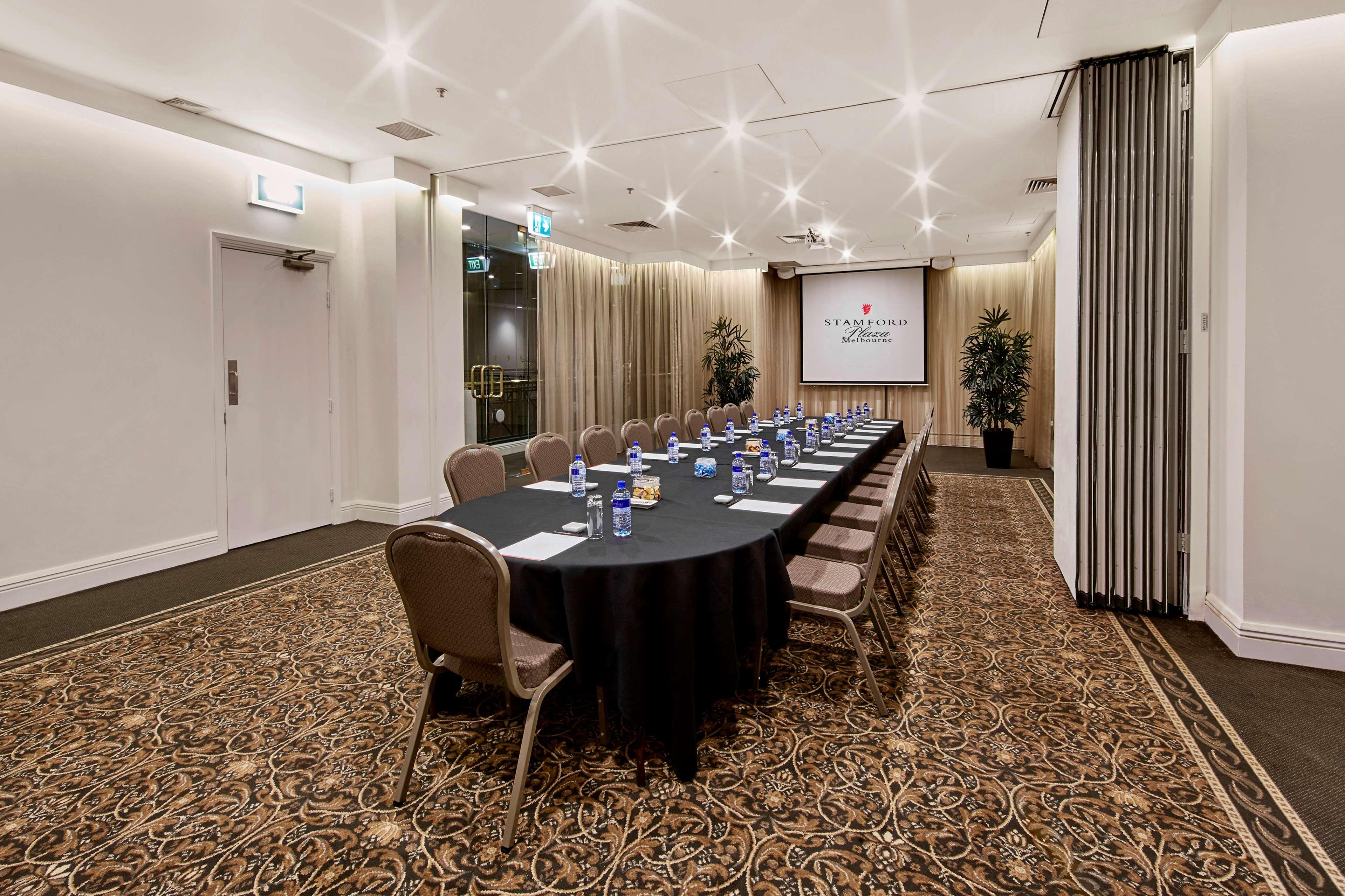 墨尔本斯坦福广场酒店 外观 照片 The photo shows a meeting room set up for a conference or presentation. There is a long, rectangular table covered with a black tablecloth, surrounded by chairs. Each seat has a water bottle placed in front of it. At the front of the room, there is a