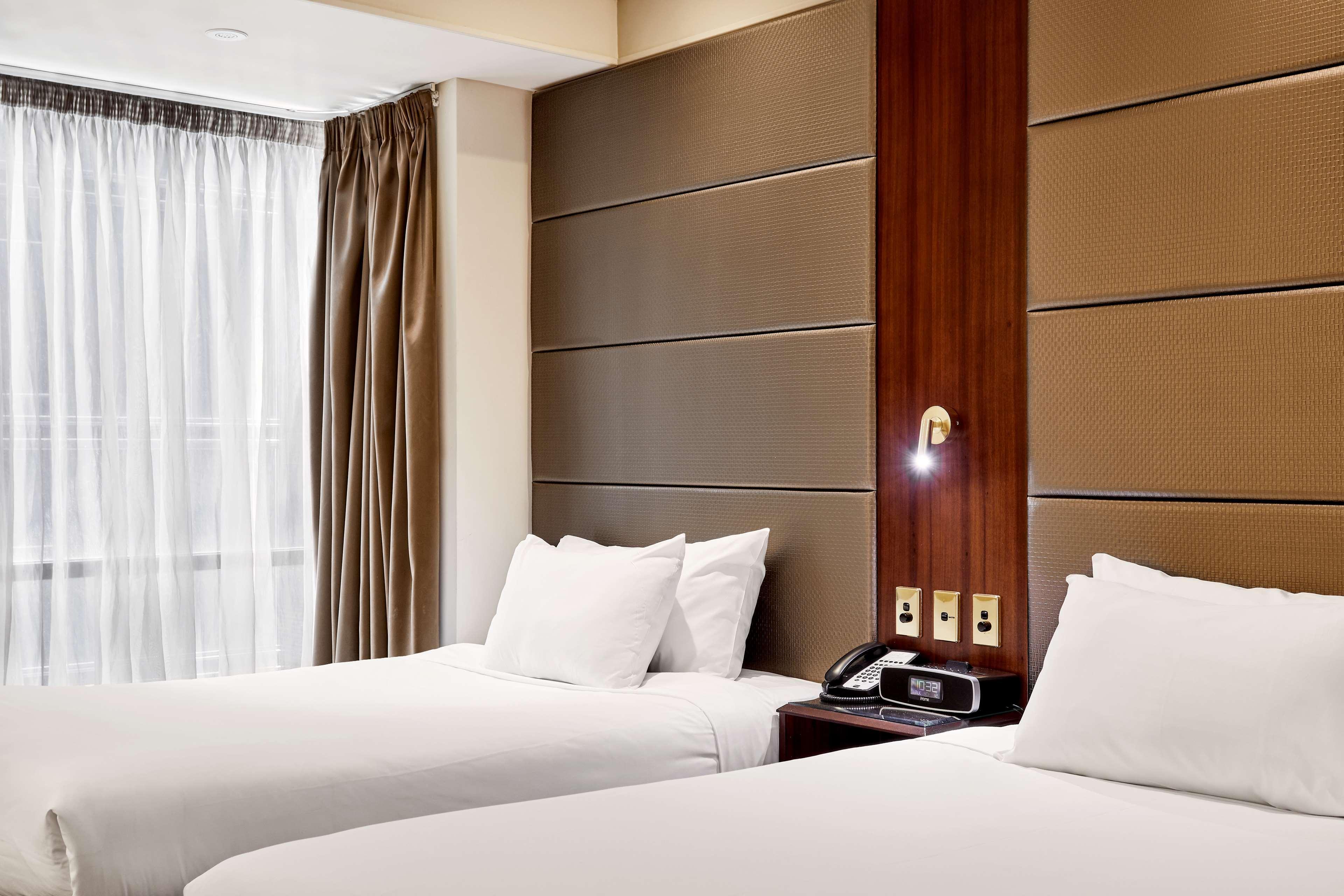 墨尔本斯坦福广场酒店 外观 照片 The photo shows a modern hotel room featuring two twin beds with white linens and fluffy pillows. The walls behind the beds have a combination of textured panels in neutral colors. There is a bedside table with a lamp and a phone. A large window with