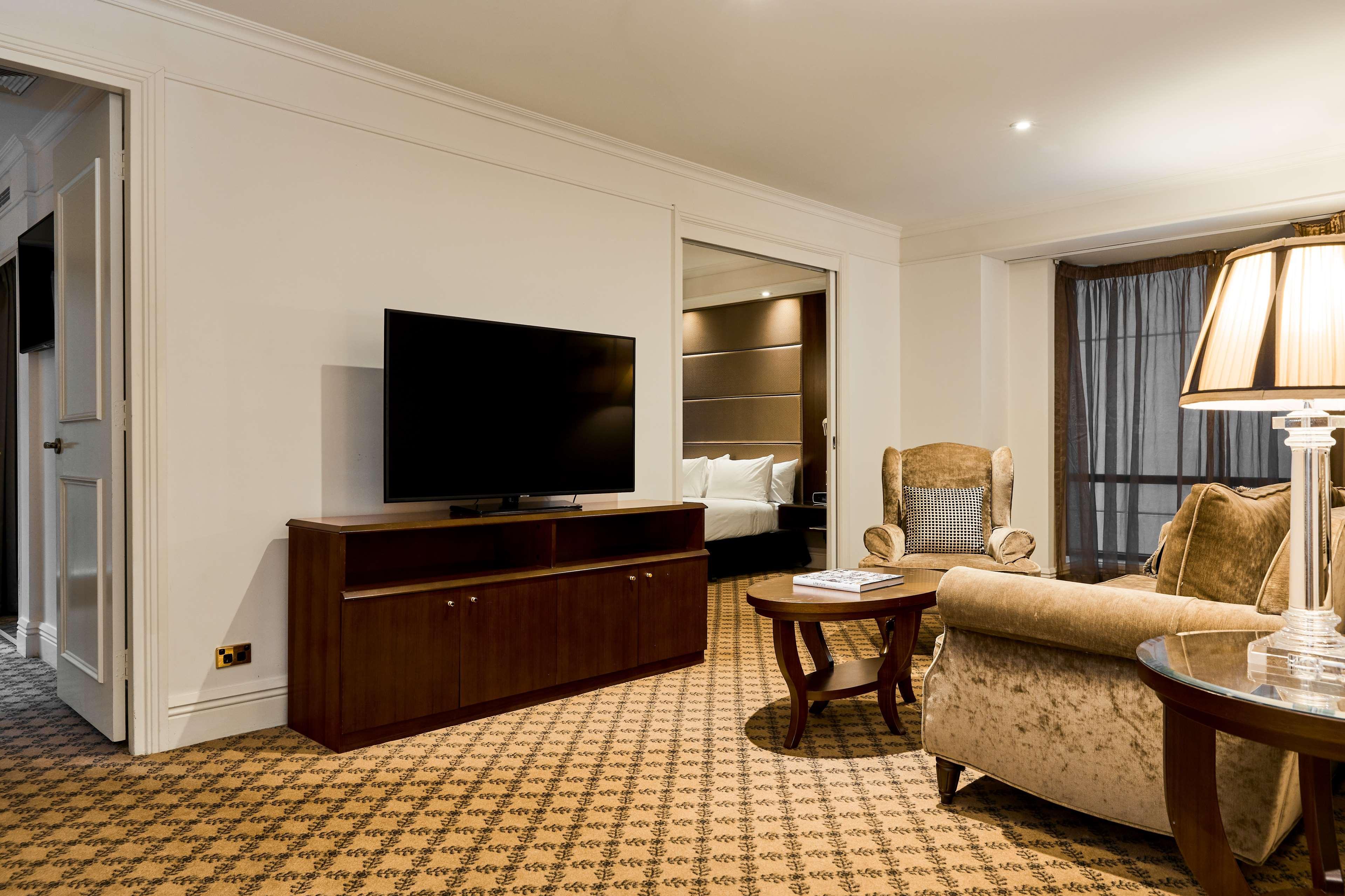 墨尔本斯坦福广场酒店 外观 照片 The photo shows a well-furnished room, likely a hotel suite or a living area. There is a large flat-screen television mounted on a wooden entertainment unit. In front of the TV, there is a round wooden coffee table. To the side, a comfortable armchai
