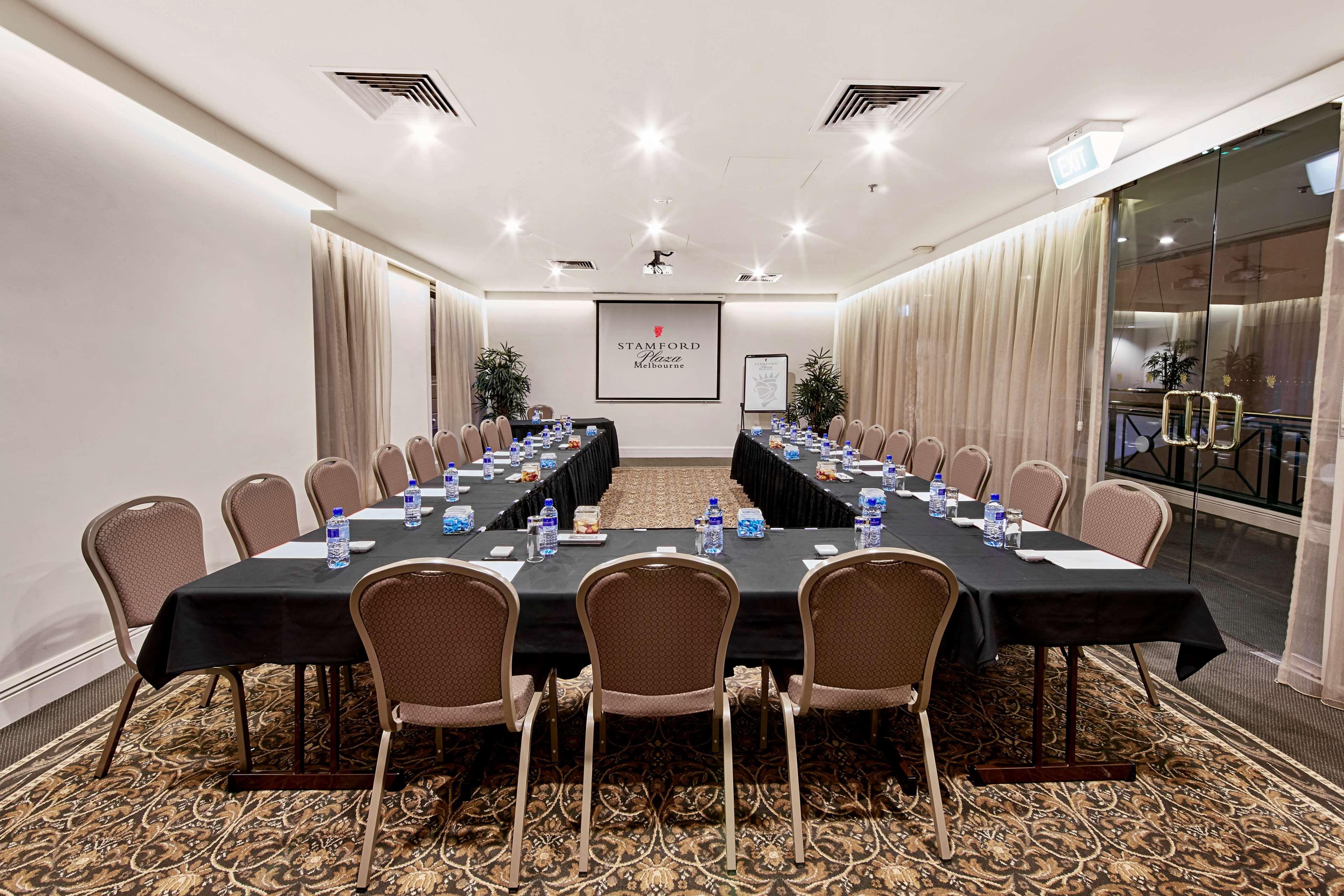 墨尔本斯坦福广场酒店 外观 照片 The photo shows a conference room set up for a meeting. There are several long tables arranged in a U-shape, covered with black tablecloths. Each table has a water bottle and notepad placed at intervals. The room is well-lit, with light-colored walls