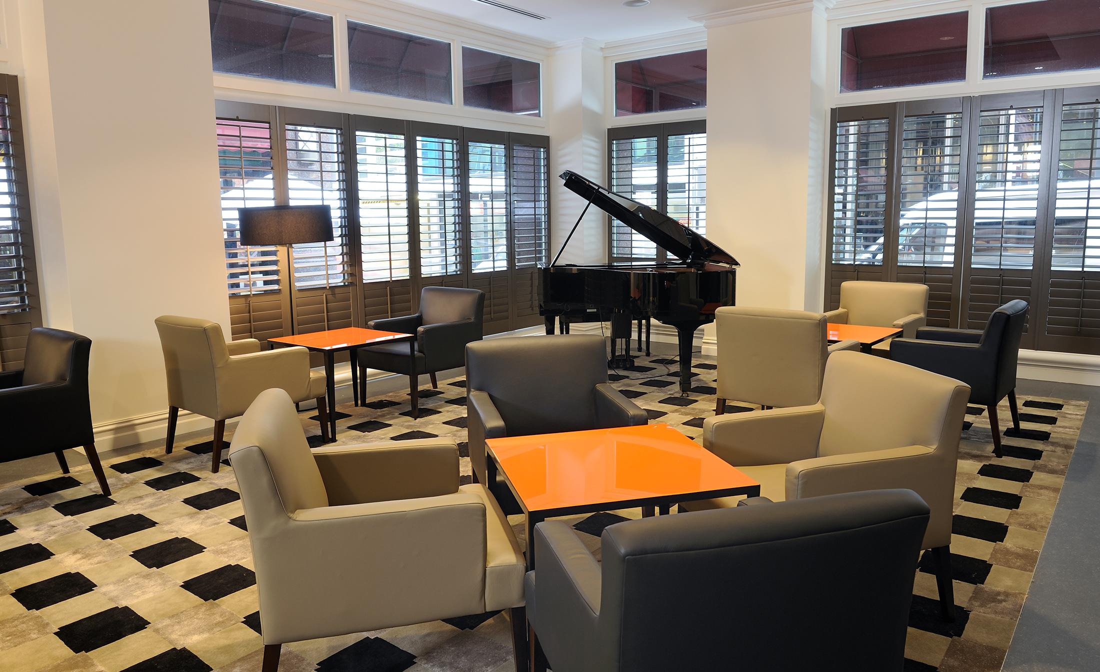 墨尔本斯坦福广场酒店 外观 照片 The photo shows a stylish indoor lounge area. It features several light gray armchairs arranged around orange square tables. In the corner, there is a black grand piano, adding an elegant touch to the space. The room has large windows with wooden shu