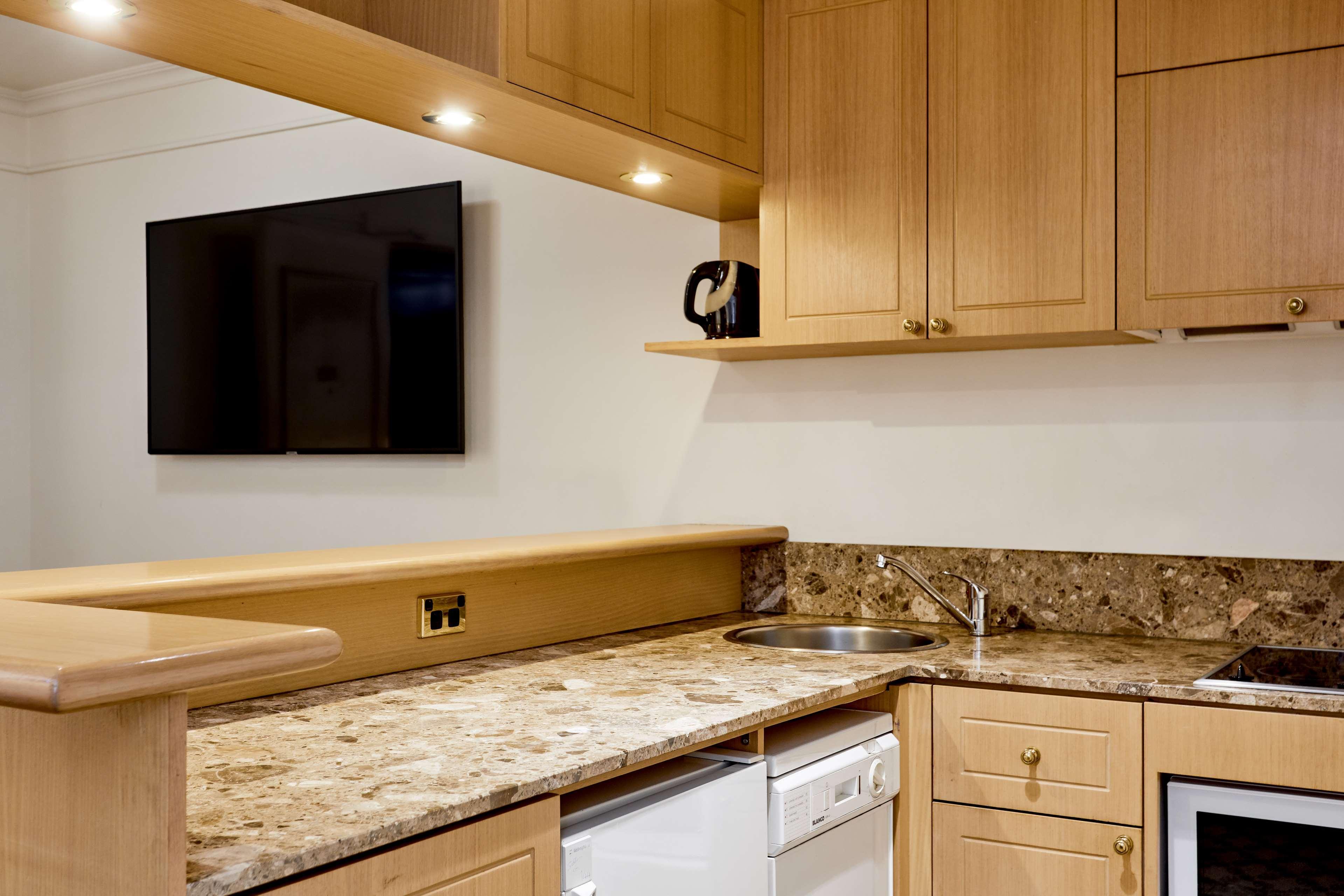 墨尔本斯坦福广场酒店 外观 照片 The photo shows a modern kitchen with wooden cabinetry and a granite countertop. There's a stainless steel sink integrated into the countertop, and appliances including a dishwasher and possibly a coffee maker on the shelf above. A flat-screen televi