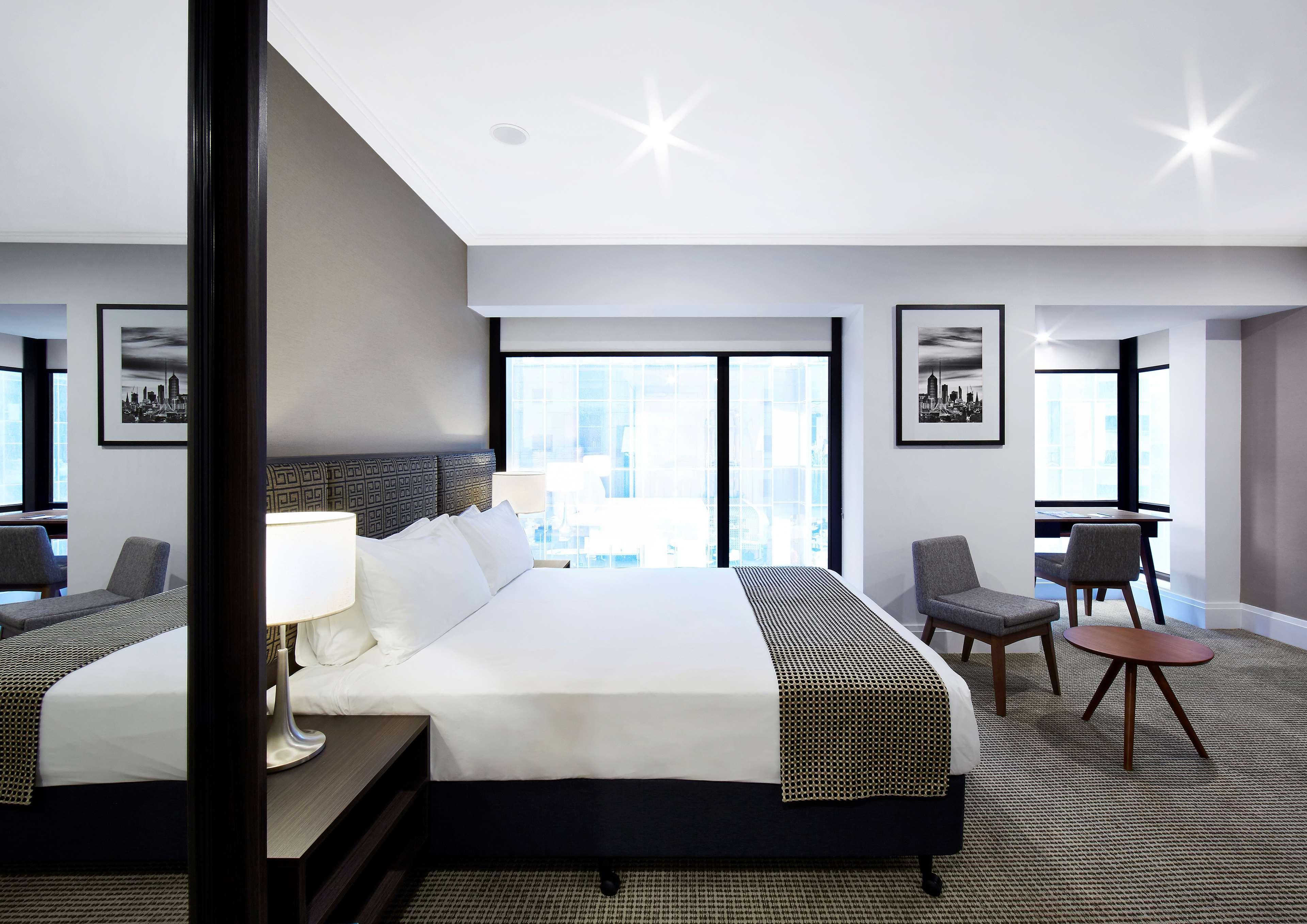 墨尔本斯坦福广场酒店 外观 照片 The image shows a modern hotel room featuring a large bed with white linens and a gray throw blanket. There are two windows that allow natural light to enter the space, and the walls are painted in a light color. In addition to the bed, there are two