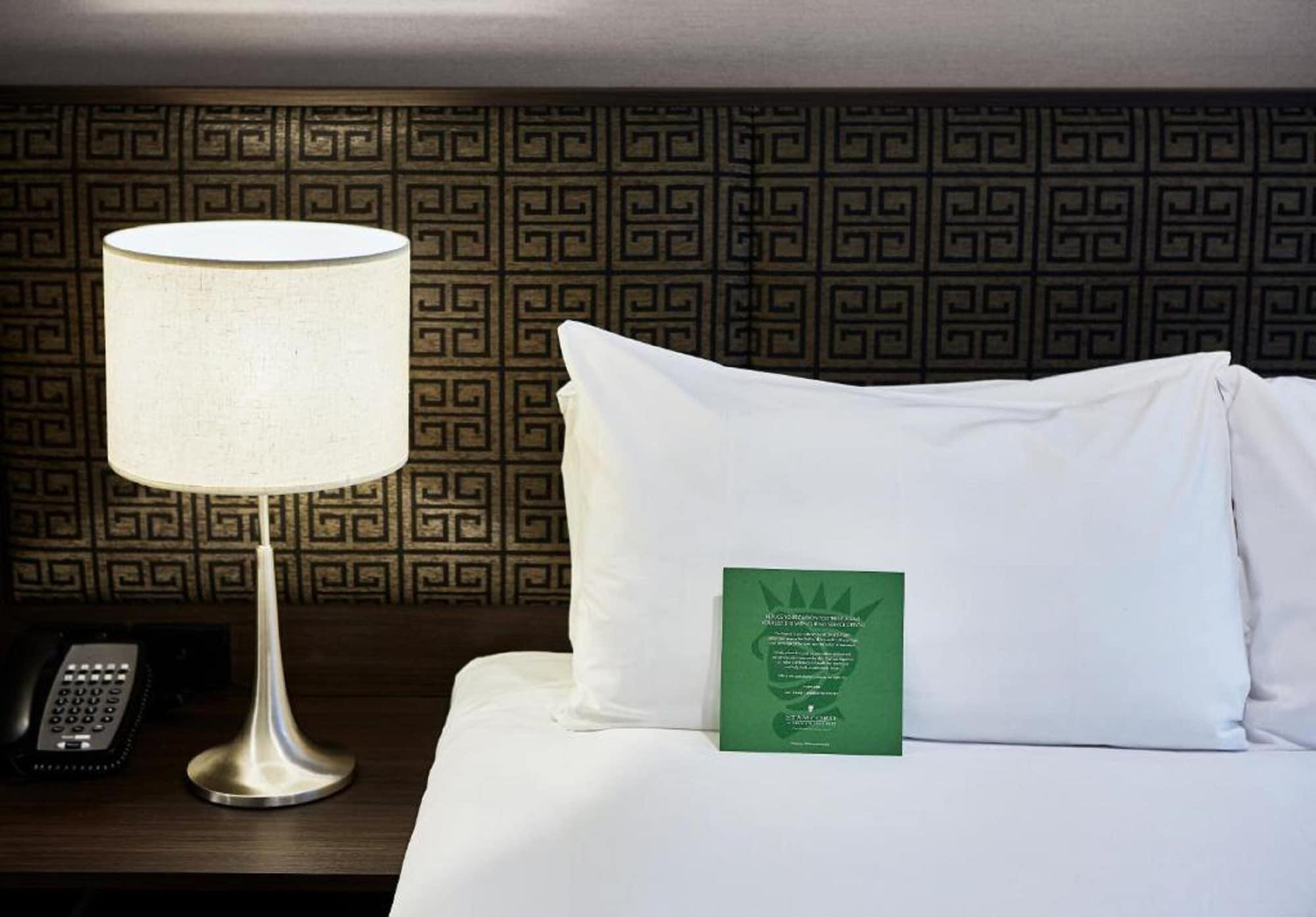 墨尔本斯坦福广场酒店 外观 照片 The photo shows a neatly made bed with a white pillow and a green item placed on top of it. Next to the bed, there is a stylish table lamp with a lampshade that appears to be white. In the background, you can see a patterned wall, which has a geometr