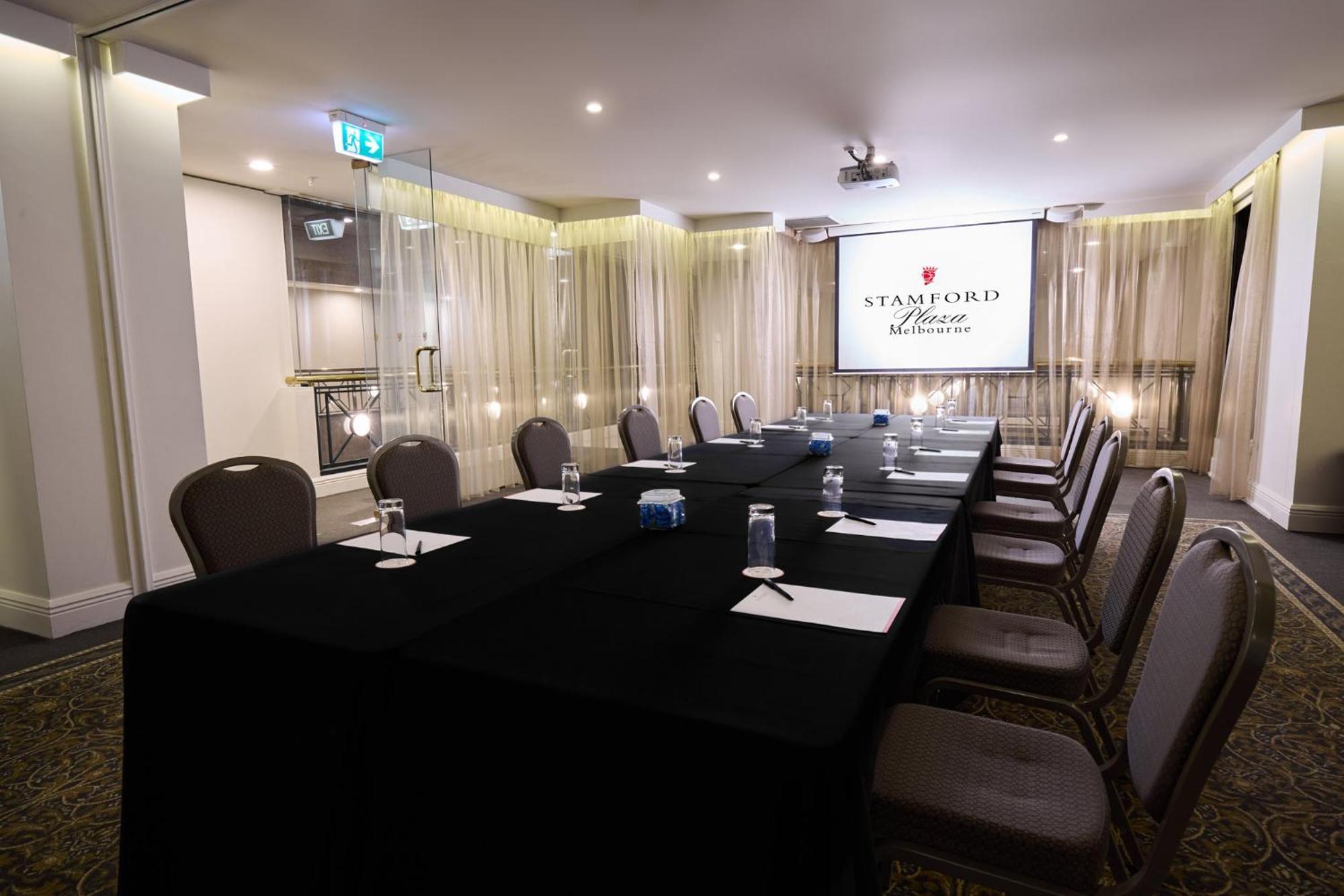 墨尔本斯坦福广场酒店 外观 照片 The photo shows a well-appointed conference room designed for meetings or presentations. There is a long rectangular table covered with a black tablecloth, surrounded by upholstered chairs. On the table, there are several folded pieces of paper and g