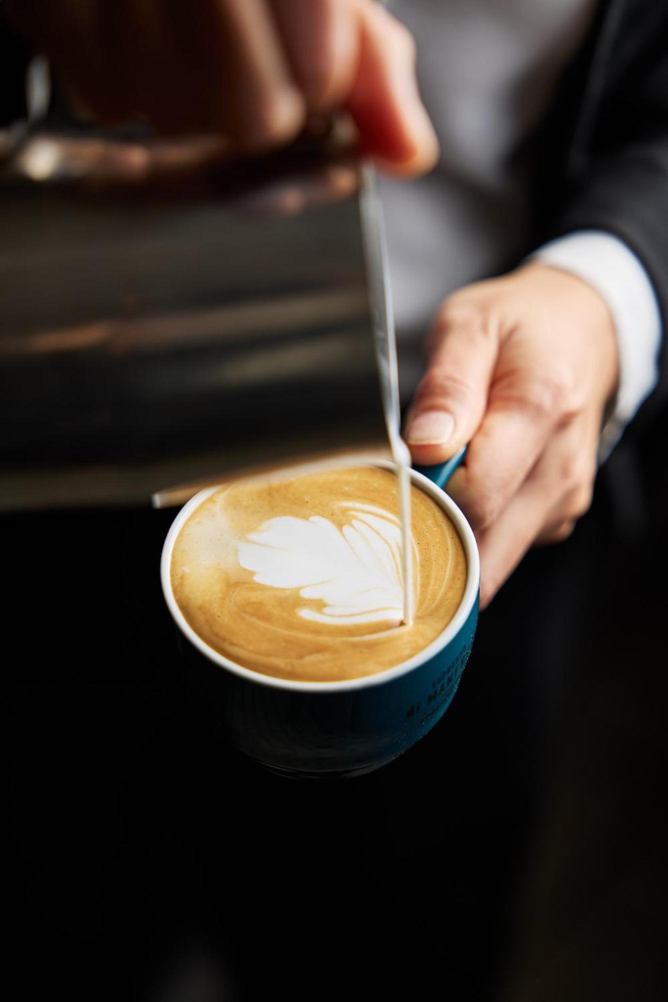 墨尔本斯坦福广场酒店 外观 照片 The photo shows a close-up of a hand holding a cup of coffee with a latte art design on the surface. The coffee appears to be a creamy, caramel color, likely a latte or cappuccino. A small stream of milk is being poured into the cup, creating a heart