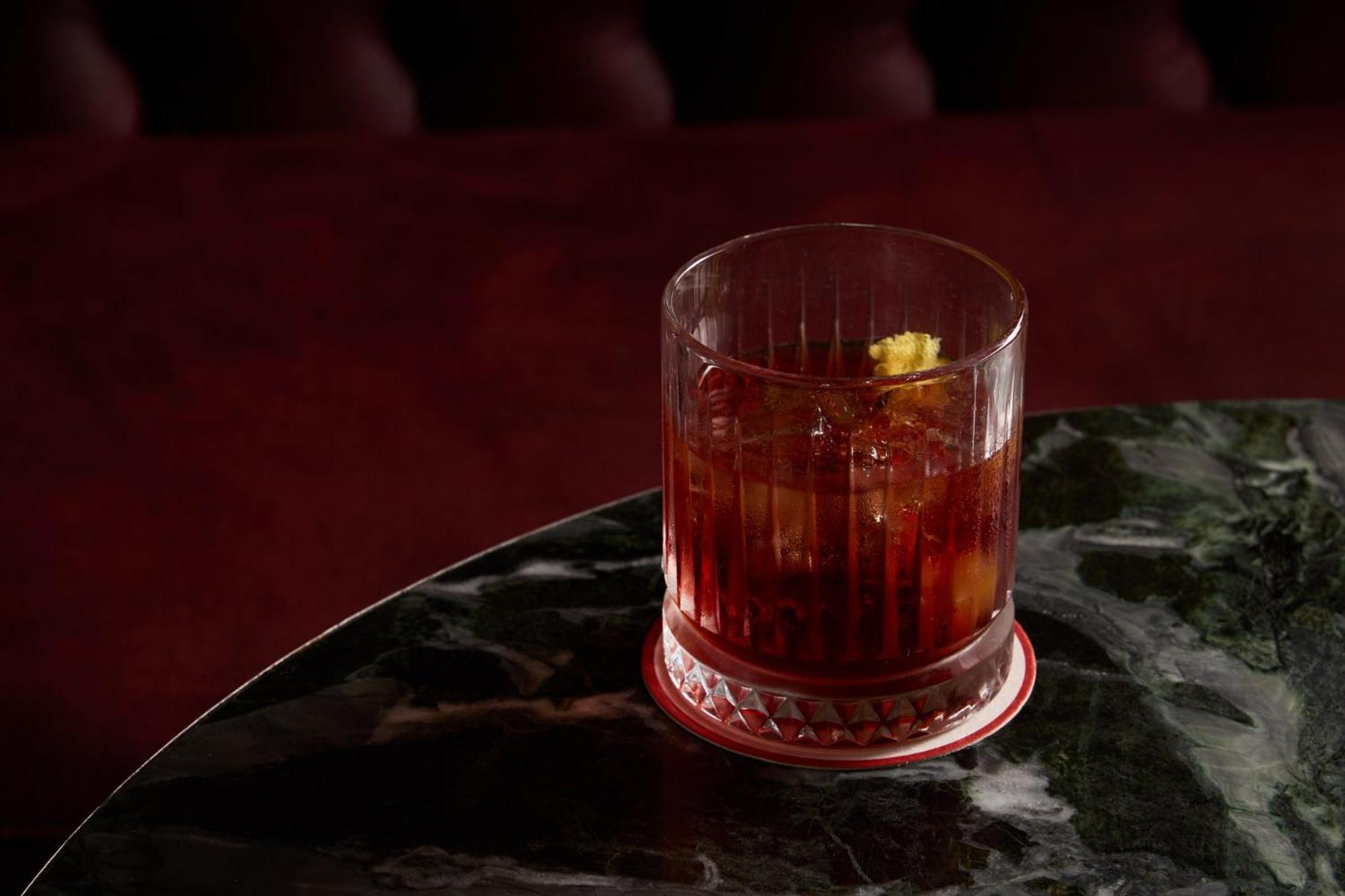 墨尔本斯坦福广场酒店 外观 照片 The photo depicts a short glass filled with a dark-colored beverage, likely an alcoholic drink. The glass has a textured design, with ice cubes visible inside. A twist of lemon peel is perched on the rim, adding a touch of garnish. The drink sits on 
