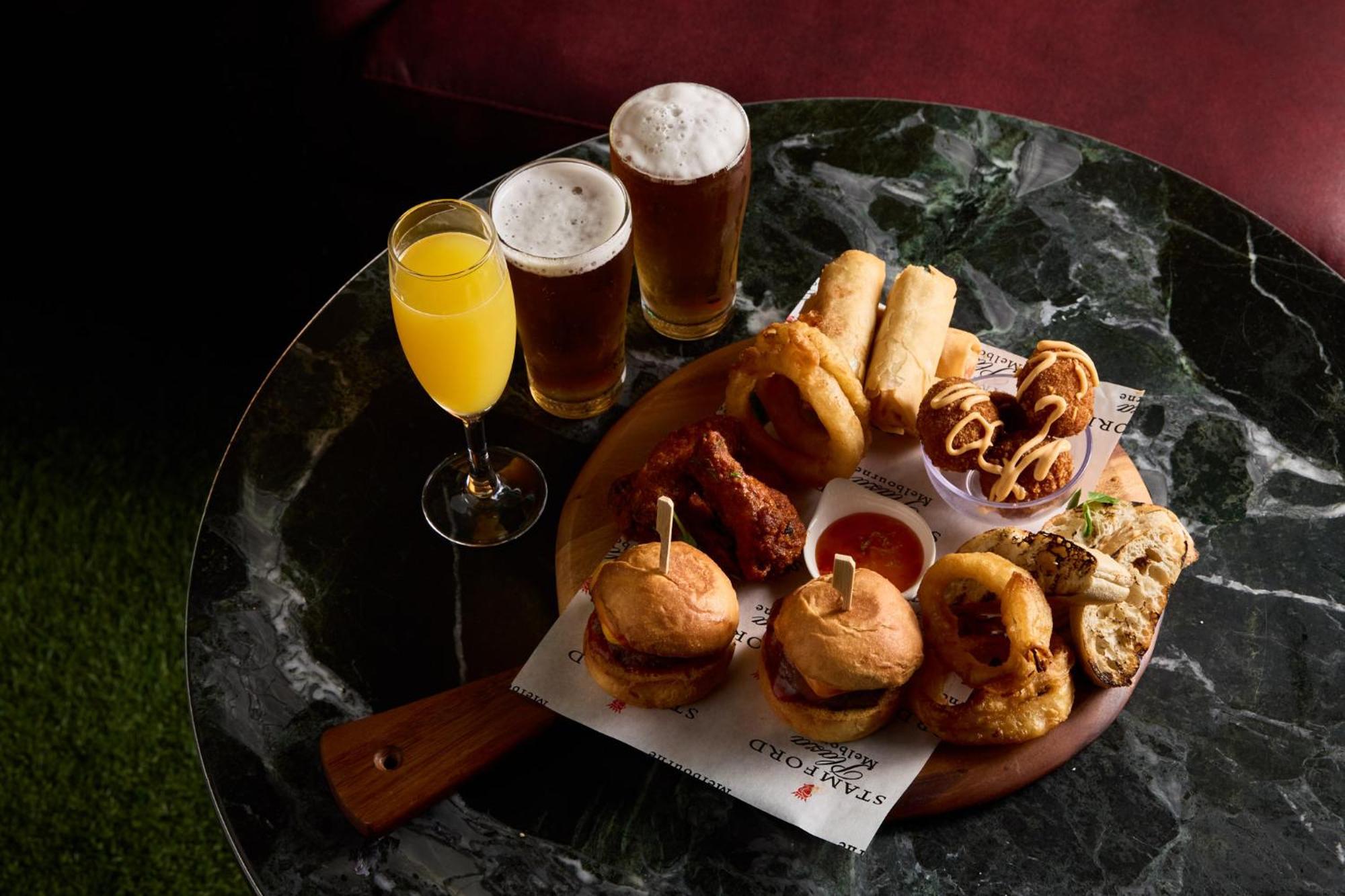 墨尔本斯坦福广场酒店 外观 照片 The photo displays a variety of food and drinks arranged on a marble table. There are small burgers garnished with skewers, crispy chicken pieces, and deep-fried snacks. Additionally, there are two glasses of beer and a cocktail, likely a mimosa, pre