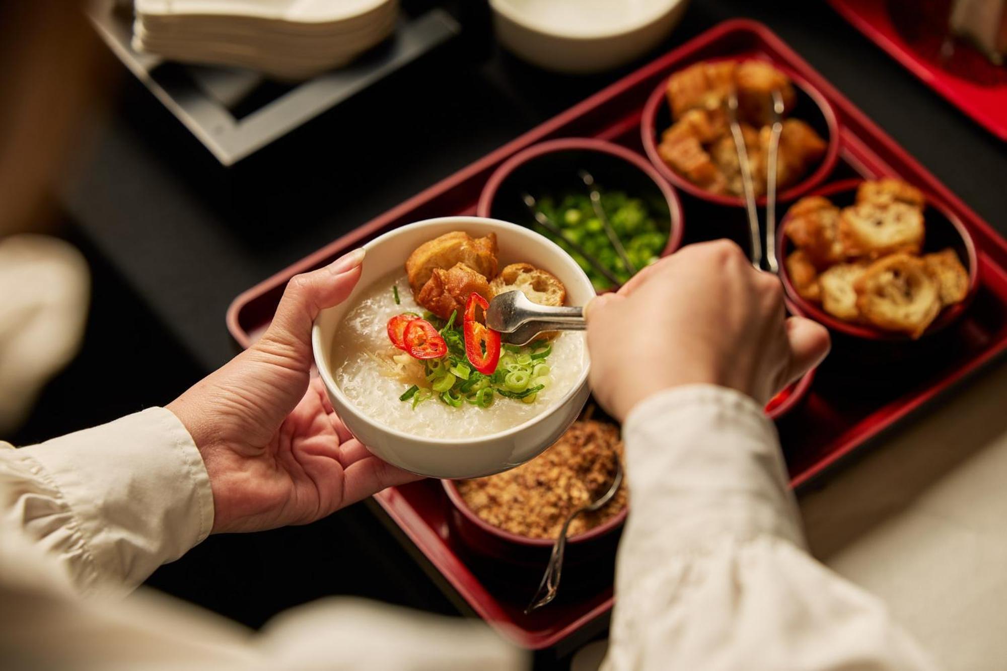 墨尔本斯坦福广场酒店 外观 照片 The photo shows a person serving food from a tray filled with various dishes. In one hand, they are holding a bowl filled with a creamy, likely rice-based dish topped with slices of chili and possibly fried tofu or other toppings. Surrounding the bow
