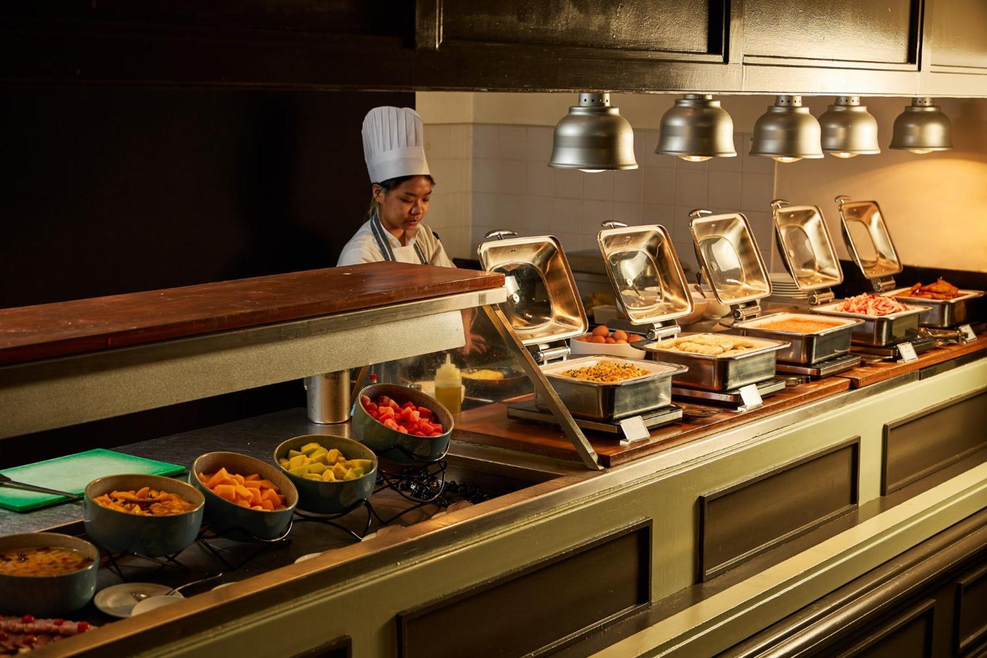 墨尔本斯坦福广场酒店 外观 照片 The photo shows a buffet setup in a dining area. There is a chef in a white uniform and hat, standing behind a counter filled with various food options. The buffet features several metal serving trays containing different dishes, with warming lights 