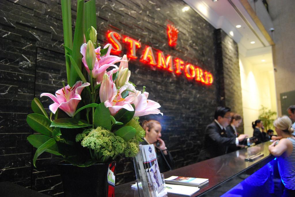墨尔本斯坦福广场酒店 外观 照片 The photo shows a hotel reception area with a prominent sign that reads "STAMFORD" in bright red letters. In the foreground, there is a decorative arrangement of flowers, primarily featuring pink lilies, along with greenery. The reception counter is 