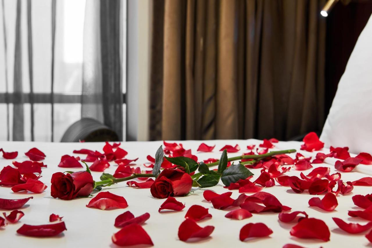 墨尔本斯坦福广场酒店 外观 照片 The photo shows a bed or a table covered with scattered red rose petals. Among the petals, there are a few whole red roses, still with their stems. The background features curtains that appear to be sheer, allowing some light to come through, and the